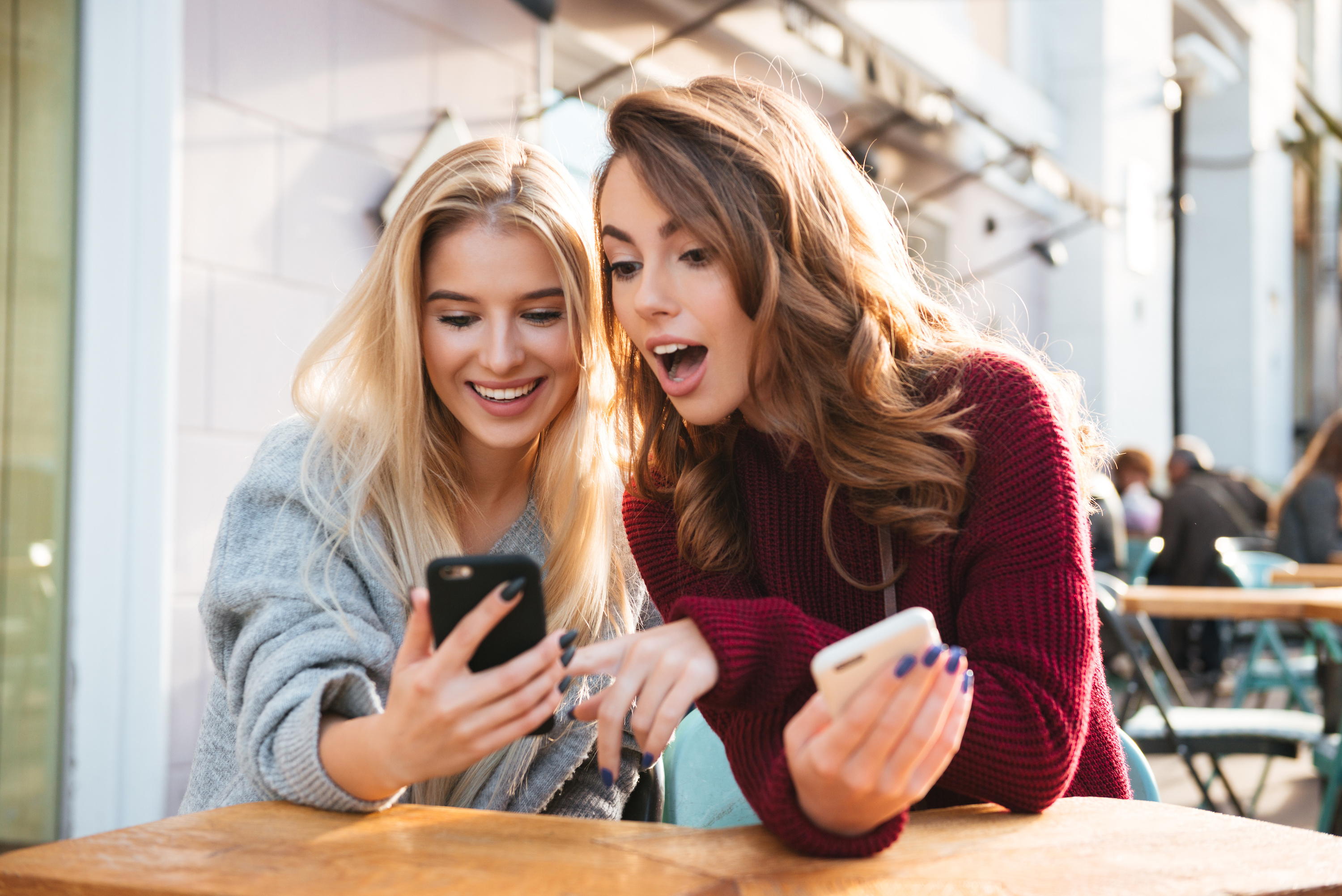 Two excited young girls using mobile phones while sitting at the cafe outdoors and pointing finger